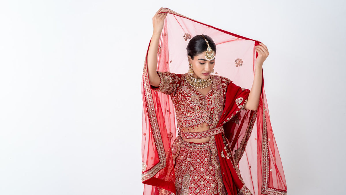 Bride in a red embroidered lehenga raising a sheer red dupatta over her head