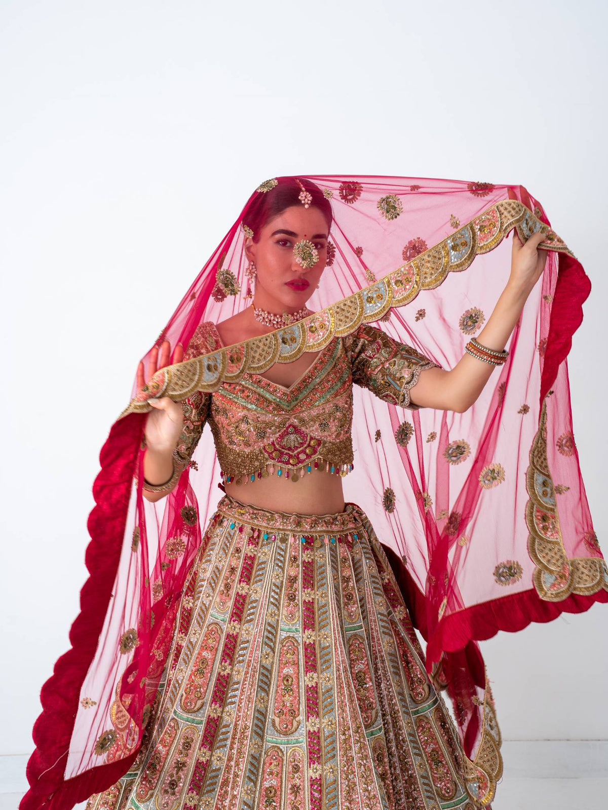 Bride holding a scalloped, embroidered red veil in a multicolour bridal lehenga