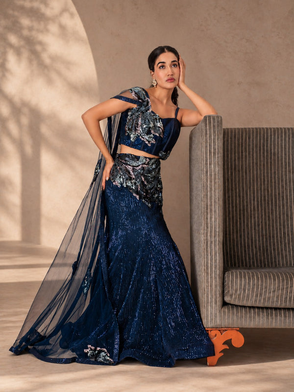 Model in the navy sequin fishtail lehenga with a sheer cape drape, beside a striped sofa